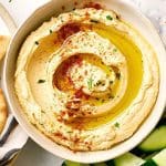 Skål med hummus toppad med olivolja och paprikakrydda, omgiven av grönsaker och kex.