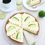 Key lime pie med grädde och limeklyftor på en glasskiva, med en bit skuren på en tallrik.