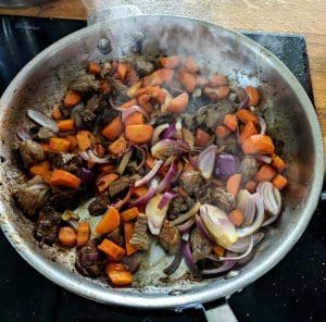 En stekpanna med kött, morötter och lök som tillagas med ånga som stiger upp.