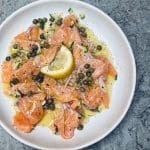 Tallrik med skivad lax, kapris, lök och citronskiva på en grå yta.