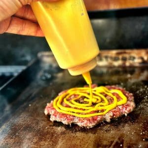 En hand som häller senap i spiralform på en köttfärsbiff som grillas.