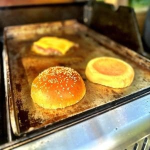 Tre hamburgare och bröd på en grillplatta, inklusive ett sesamfröbröd.