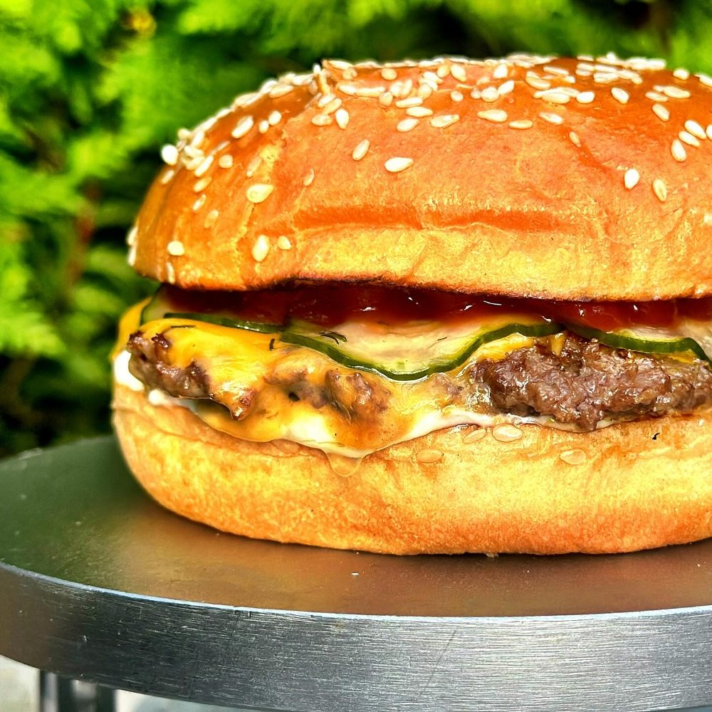 Kolbulle 11 Hamburgare med sesamfröbröd, ost, kött och gurka på en metallplatta.