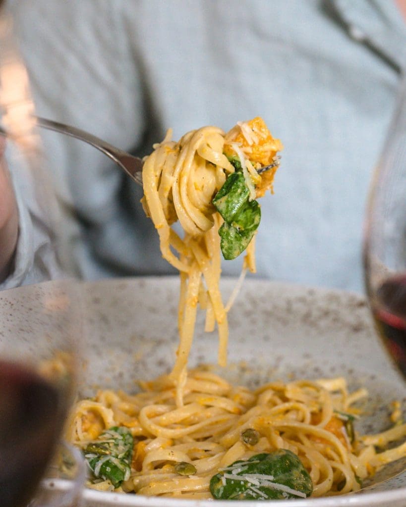 Hannas krämiga salsicciapasta 9 En gaffel med pasta, butternutpumpa och spenat i en skål, med vin i bakgrunden.