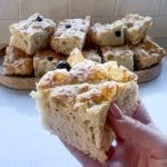En hand som håller en bit focaccia bröd med oliver, med fler bitar i bakgrunden.