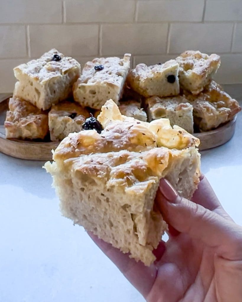 Naanbröd 9 En hand som håller en bit focaccia bröd med oliver, med fler bitar i bakgrunden.