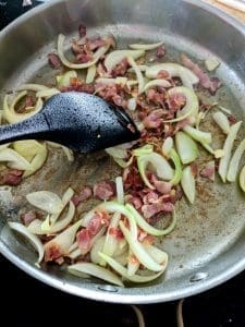 Stekt lök och bacon som tillagas i en metallpanna med en svart stekspade.