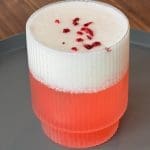 En drink med lager av rosa vätska och vit skum, dekorerad med torkade hallon på toppen.