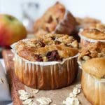 Äppelmuffins med smuldeg på en träskärbräda, med äpple och havregryn i bakgrunden.