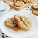 Tre chokladchip-kakor på en vit tallrik med fler kakor i bakgrunden.