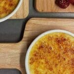 Två skålar med crème brûlée på en träskärbräda med bär i bakgrunden.