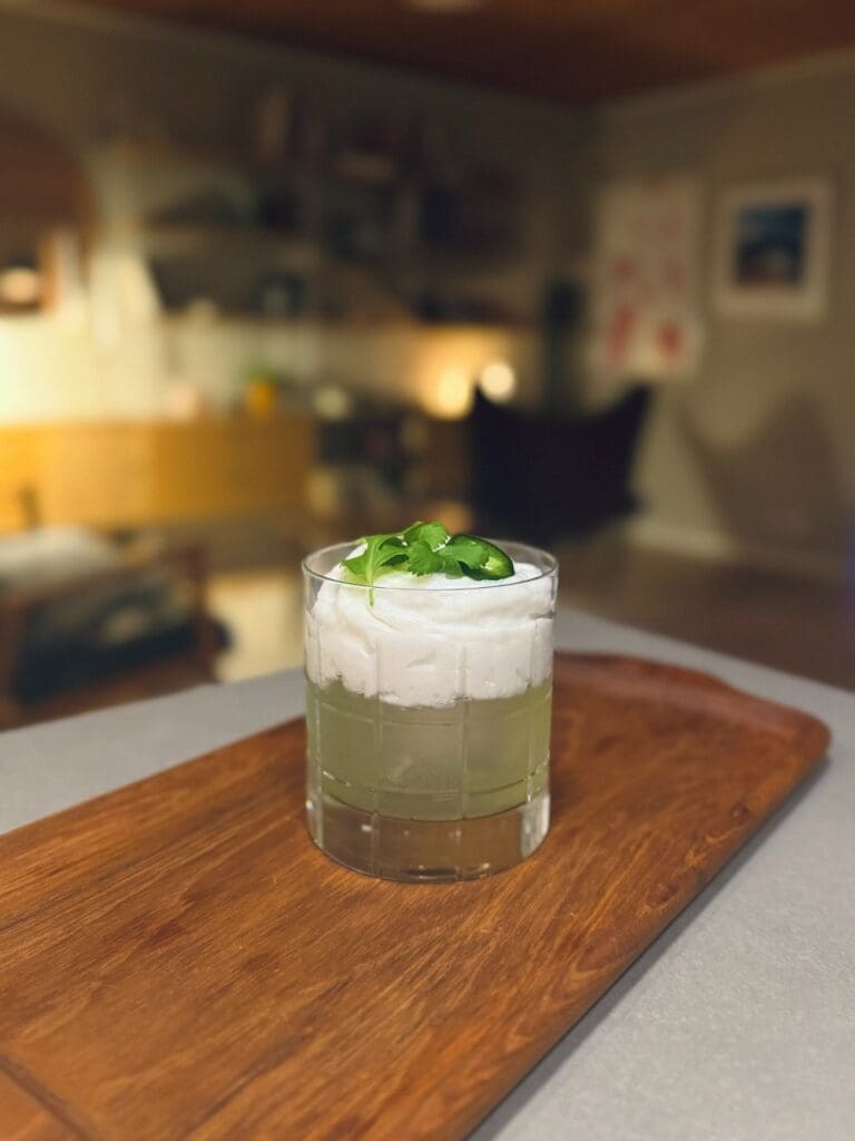 Amaretto sour 10 En drink i ett glas med kokosgrädde och koriander på toppen, placerad på en träskärbräda.