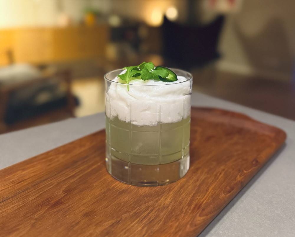 Rhubarb exotic 7 En drink med koriander och kokosgrädde i ett fyrkantigt glas på en träskärbräda.
