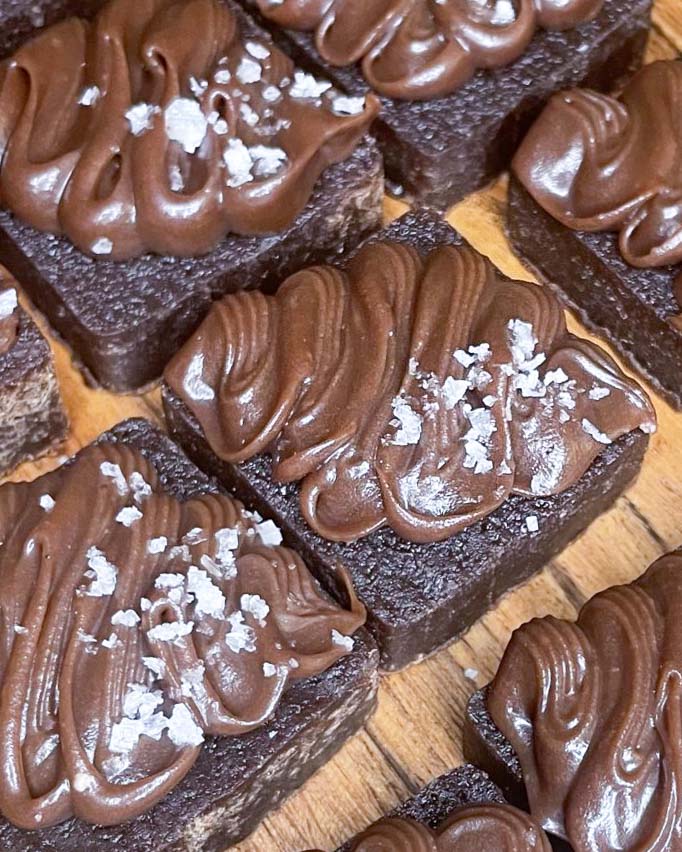 Hasselnötbrownie med nutellafrosting 10 Brownies med chokladglasyr och havssalt på en träyta.