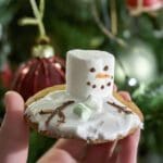 En hand som håller en kaka med en smält snögubbe gjord av marshmallow och glasyr.