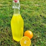 Flaska med gul apelsinsaft bredvid en hel och en halverad apelsin på gräs.