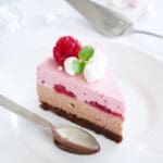 En bit cheesecake med hallon och choklad, dekorerad med hallon och grädde på en tallrik.