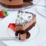 En bit kärleksmums cheesecake med chokladtopping och kokosflingor på en vit tallrik.