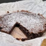 En kladdkaka med pudrat socker och en bit skuren ut, placerad på bakpapper.