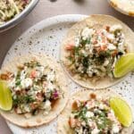 Tre lax tacos med grönsaker, sås och limeklyftor på en tallrik.