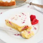 En bit mazarinkladdkaka med grädde och hallon på en vit tallrik med gaffel.