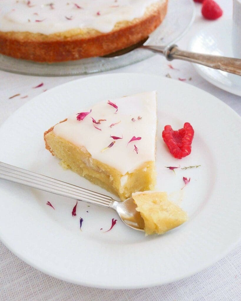 Kladdkaka med vaniljswirl och jordgubb 9 En bit mazarinkladdkaka med glasyr och blommor på en vit tallrik med gaffel.
