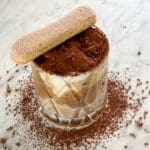 Ett glas med tiramisu latte toppat med kakao och en savoiardi-kaka ovanpå.