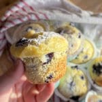 En person håller en blåbärsmuffin med vaniljkräm i handen, med flera muffins i bakgrunden.