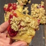 En hand som håller en bit hallonpajblondie med havregrynstopping och vaniljkräm.