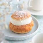 En semla med grädde på en ljusblå tallrik, med pudrat socker på toppen.