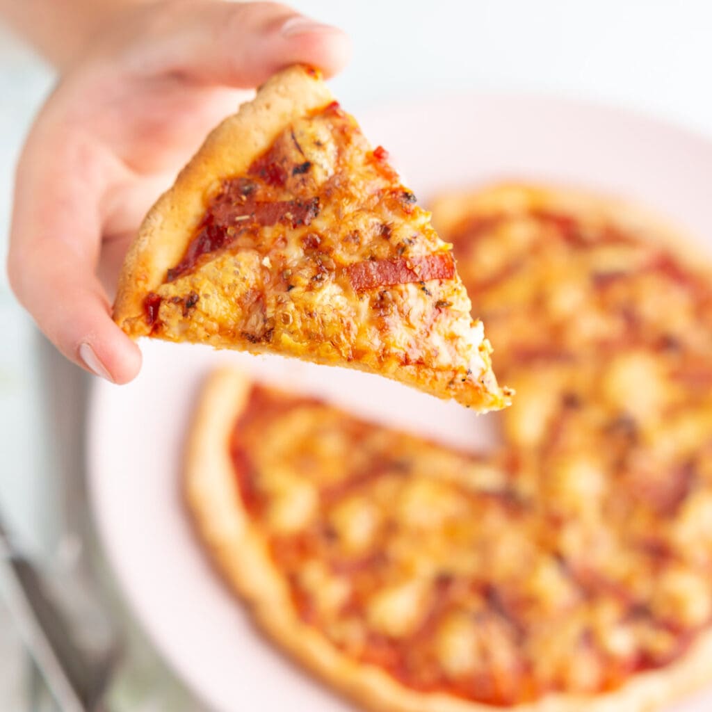 Pizza på västerbottensost och räkor 8 En hand som håller en skiva pizza med pepperoni och ost framför en tallrik med fler pizzaskivor.