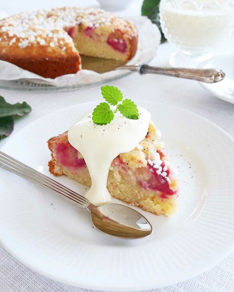 Saftig rabarberkaka med toscatäcke 9 En bit rabarberkaka med kardemumma serverad med grädde och en mintkvist på en tallrik.