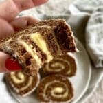 En hand som håller en skiva chokladrulltårta med vaniljfyllning, med flera skivor på en tallrik i bakgrunden.