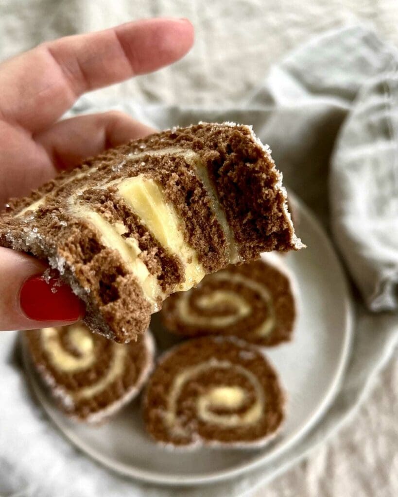 Morotsrulltårta med frosting på brynt smör 9 En hand som håller en skiva chokladrulltårta med vaniljfyllning, med flera skivor på en tallrik i bakgrunden.