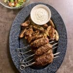 Grillade shish kebabspett serverade med vitlökssås och ugnsrostad potatis på en blå tallrik.