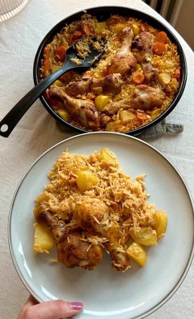 Birijan 6 En portion kyckling med ris och grönsaker på en tallrik, med en gryta i bakgrunden.