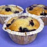 Tre blåbärsmuffins i pappersformar på en lila bakgrund.