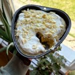 En hjärtformad citronkladdkaka med vit choklad och grädde, hålls i handen vid ett fönster.