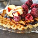Glutenfria våfflor toppade med skivor av banan, hallon och chokladsås på en tallrik.