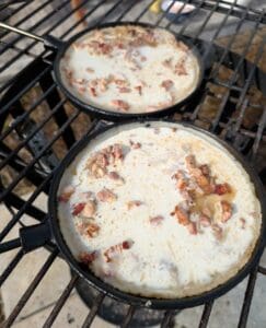 Två stekpannor med kolbulle och bacon som tillagas på en grill.