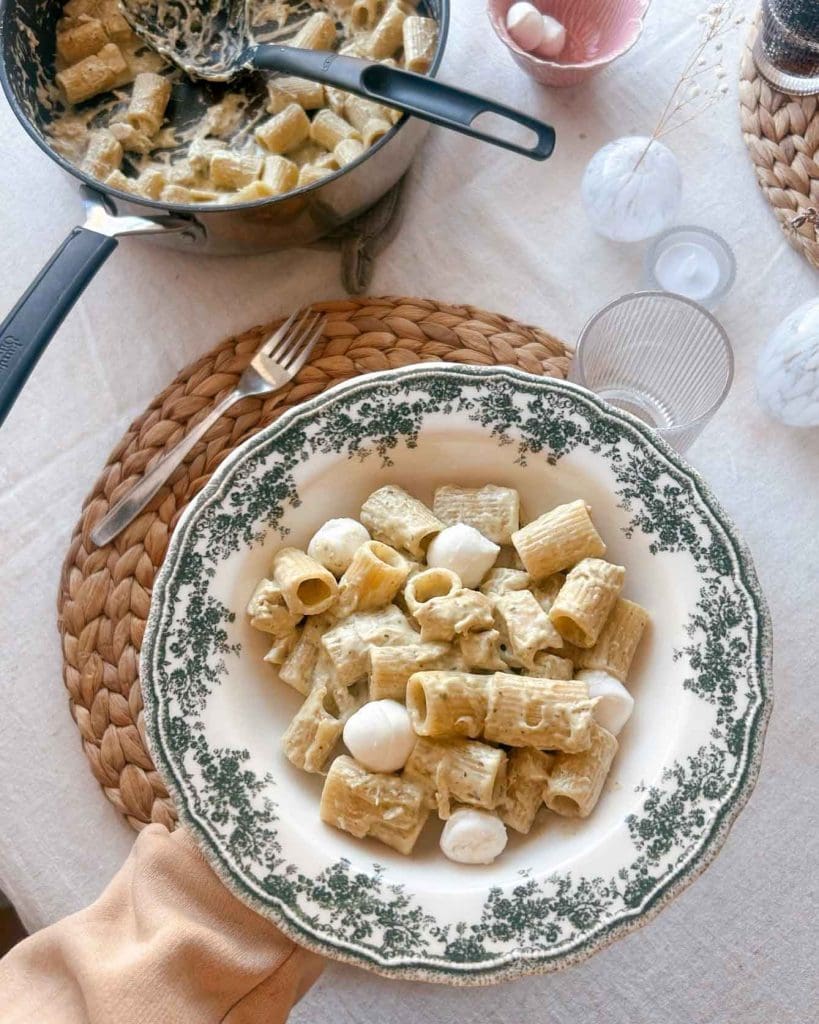Tomatsås med soltorkad tomat och riven fetaost 9 Tallrik med pasta, pesto och mozzarella bollar, serverad med en kastrull i bakgrunden.