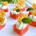 Vattenmelonbitar toppade med fetaostkräm och krispiga örter på en vit tallrik.